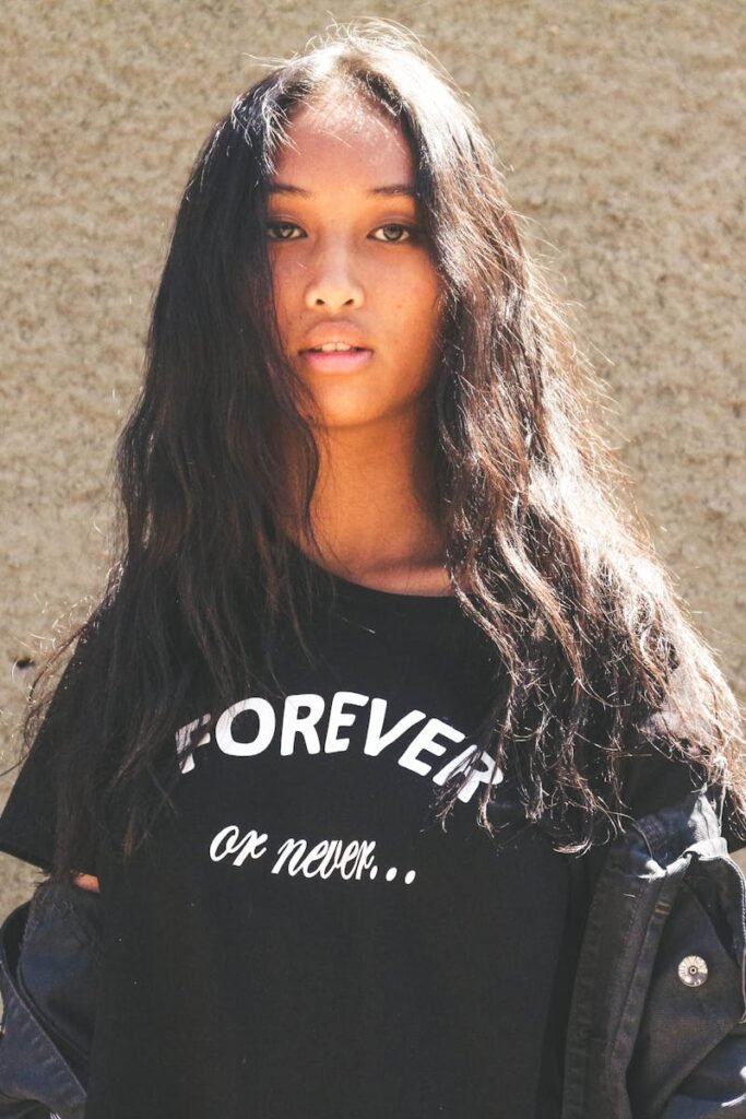 pexels-photo-1251171-1251171 Portrait of a young woman with long hair posing against a textured wall, wearing a black t-shirt in sunlight.