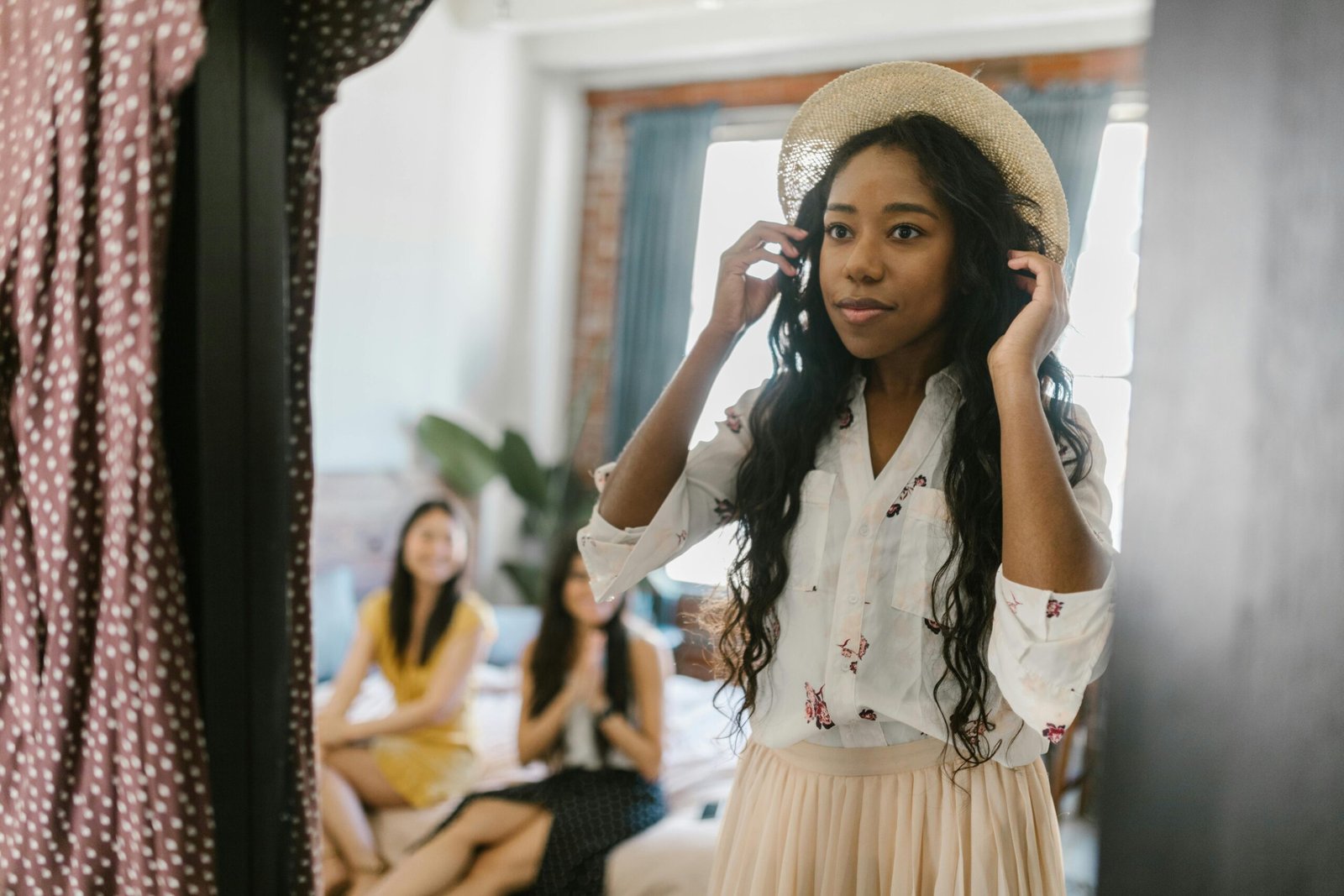 Stylish young woman trying on a hat in front of a mirror with friends in background.